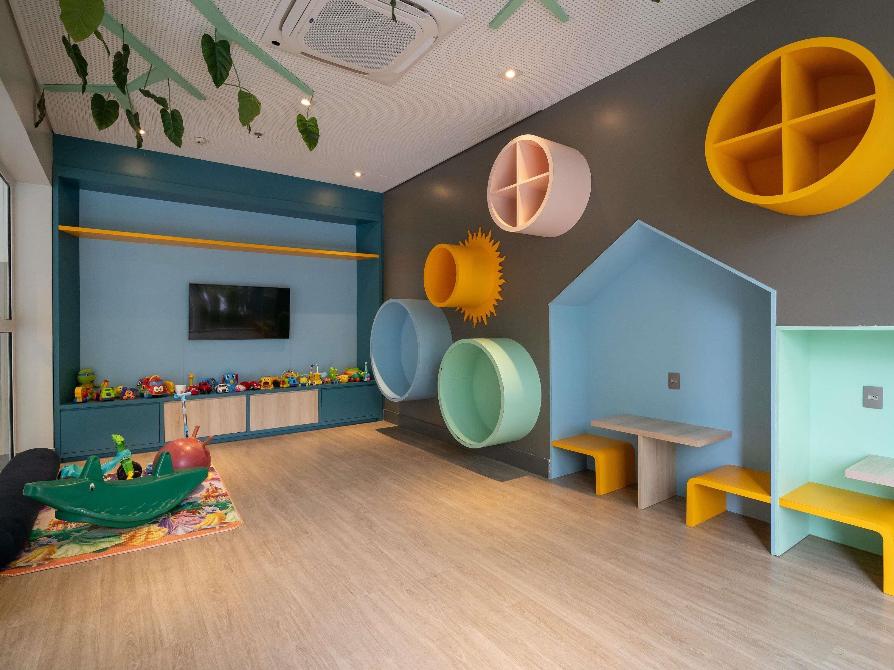 Un área de juegos infantiles con mobiliario colorido, un televisor montado y plantas colgantes.