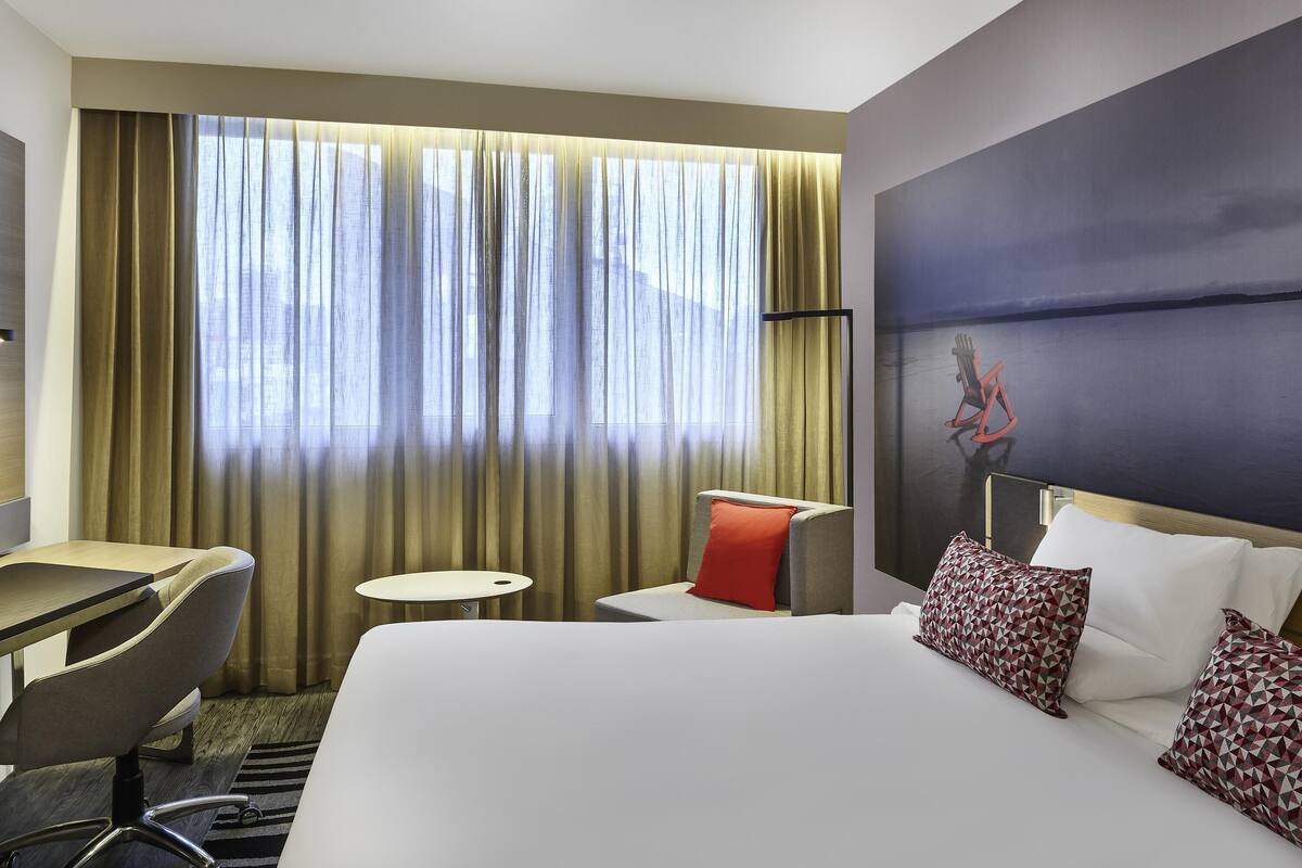 Een hotelkamer met een bed, bureau, stoel, klein tafeltje en een groot raam met gordijnen.