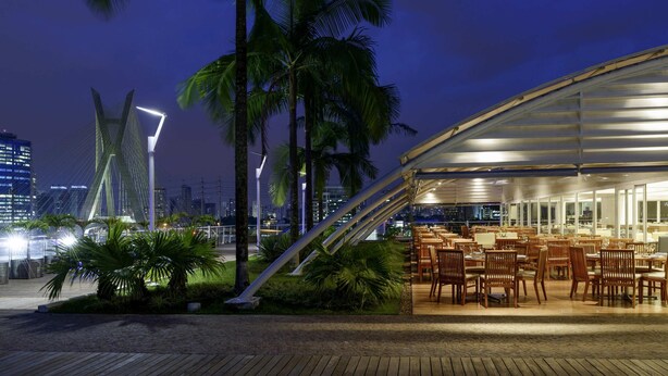 Een restaurant aan het water met terras, palmbomen en een stadsachtergrond.