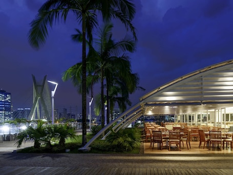 A waterfront restaurant with outdoor seating, palm trees, and a modern bridge in the background.. Novotel São Paulo Morumbi