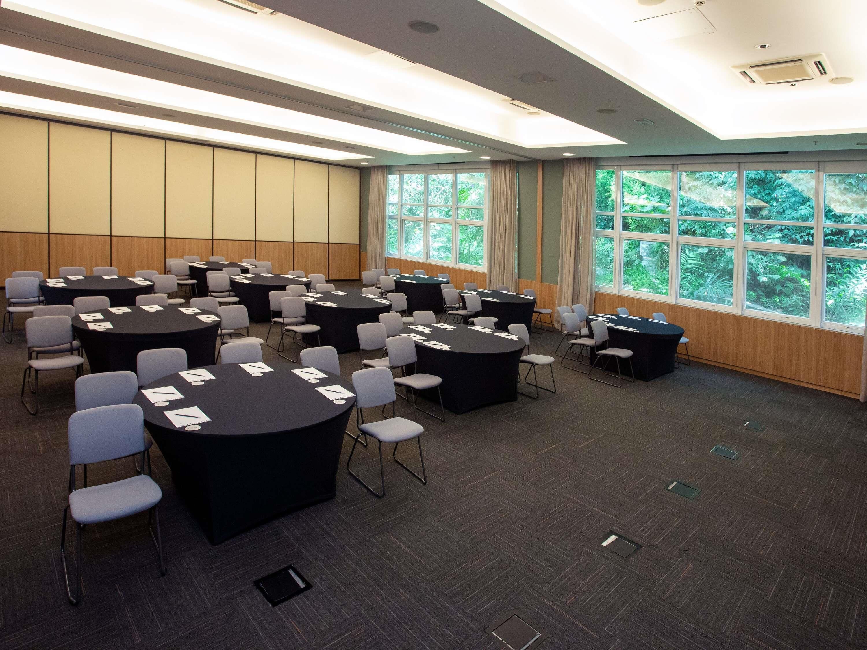 A conference room with multiple round tables, chairs, and large windows with a view of greenery.