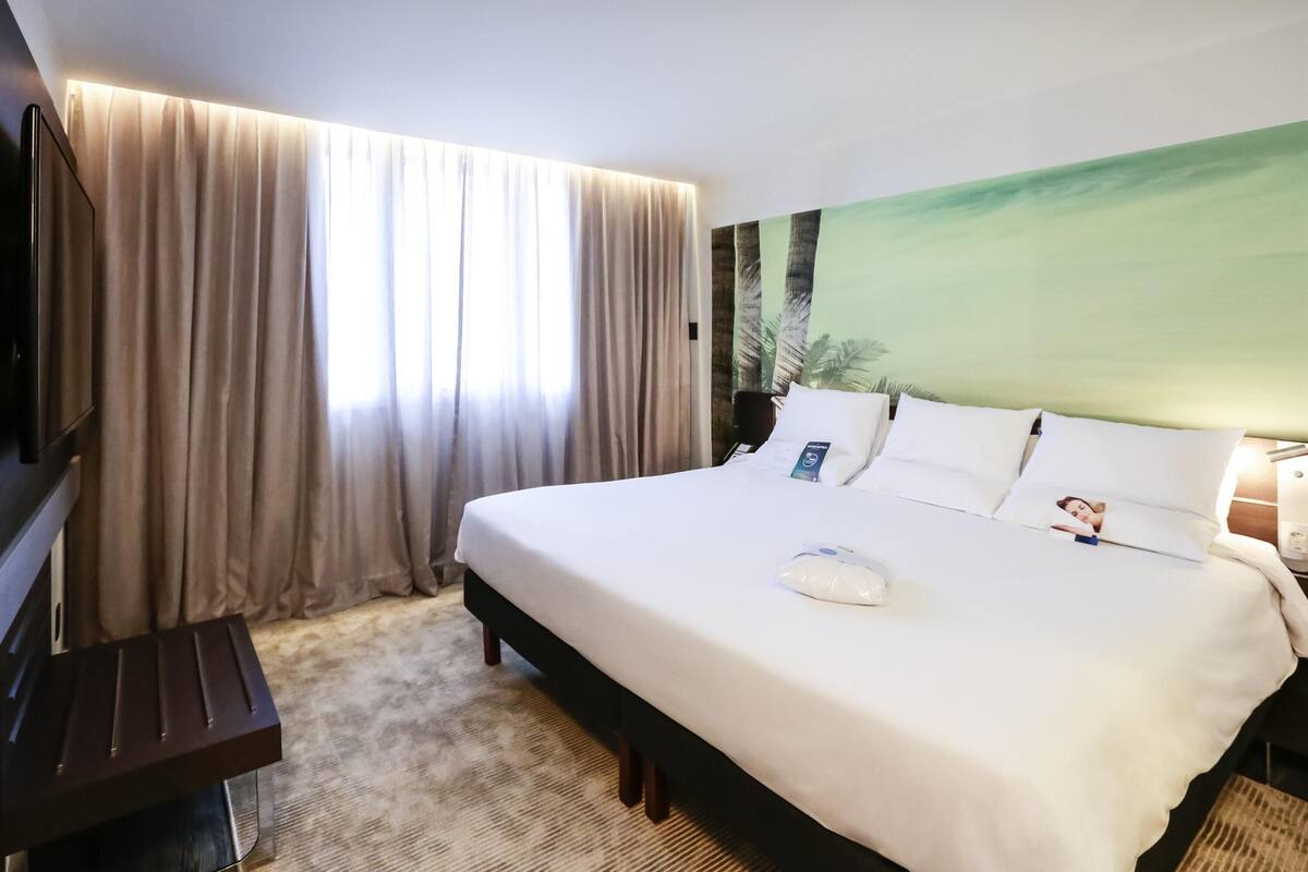 Una habitación de hotel con cama, cortinas, televisor y un cuadro en la pared.