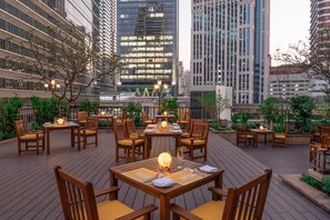 Un espace de restauration en plein air, avec des tables et des chaises en bois, entouré de verdure et offrant une vue sur un paysage urbain avec des bâtiments modernes.
