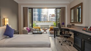 Habitación de hotel con cama, escritorio, silla, mesita pequeña y balcón con plantas y vistas a la ciudad.