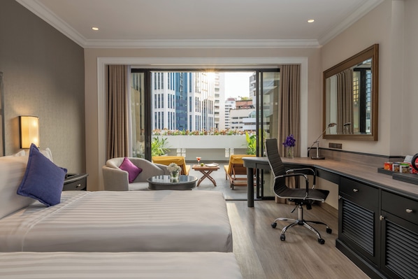 Habitación de hotel con cama, escritorio, silla, mesita pequeña y balcón con plantas y vistas a la ciudad.