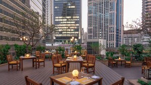 Un espacio de comedor al aire libre con mesas y sillas de madera, rodeado de follaje y edificios de la ciudad.