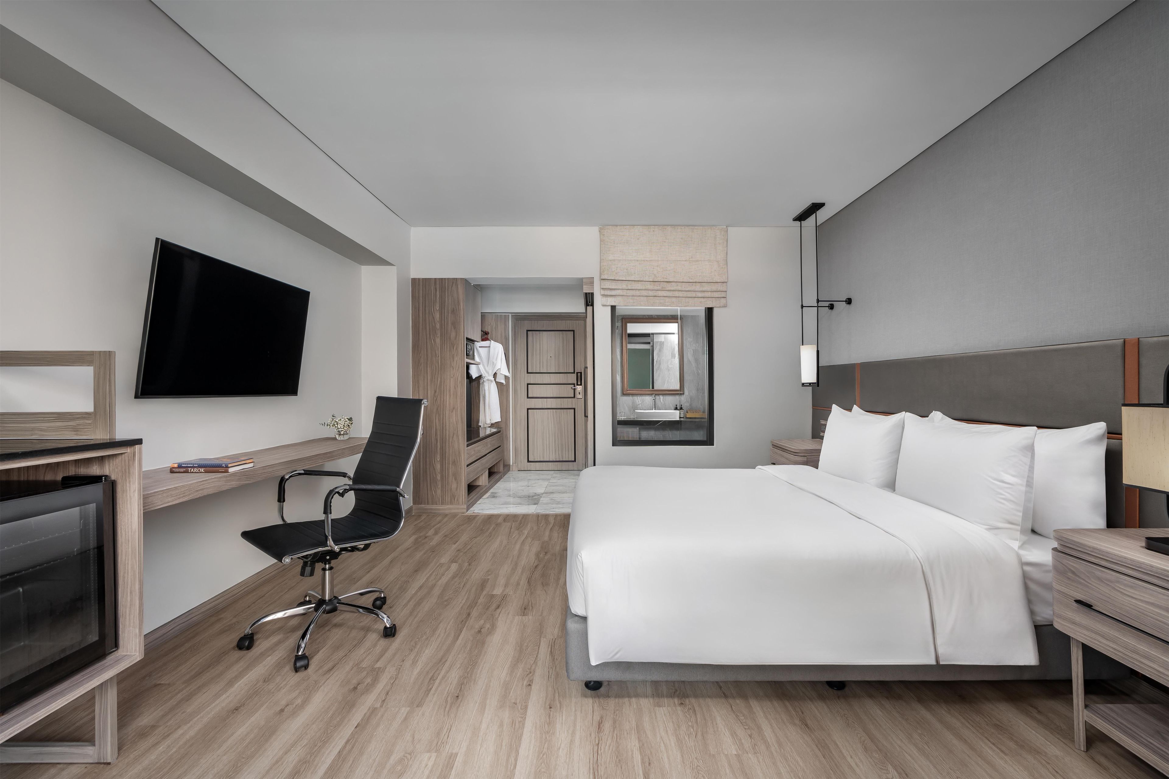 A modern hotel room with a flat-screen TV, a desk, a chair, a bed with white bedding, and a bathroom visible through an open door.