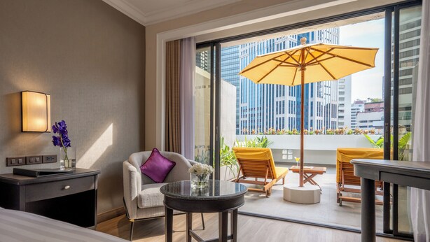 A modern hotel room with a large window, a round table, a sofa, and a small desk with a lamp.