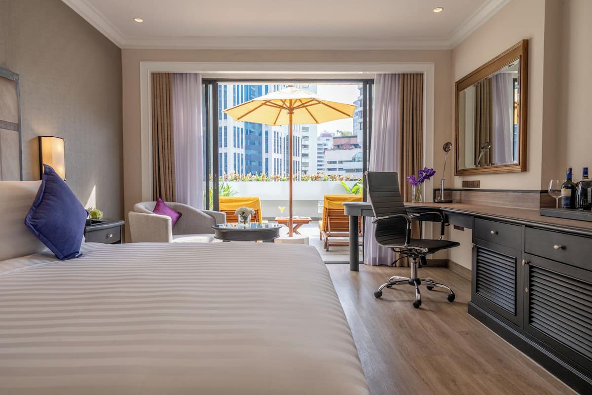 Une chambre d’hôtel avec un grand lit, un bureau avec une chaise, une petite table avec un vase, une grande fenêtre avec des rideaux et un balcon agrémenté de plantes.