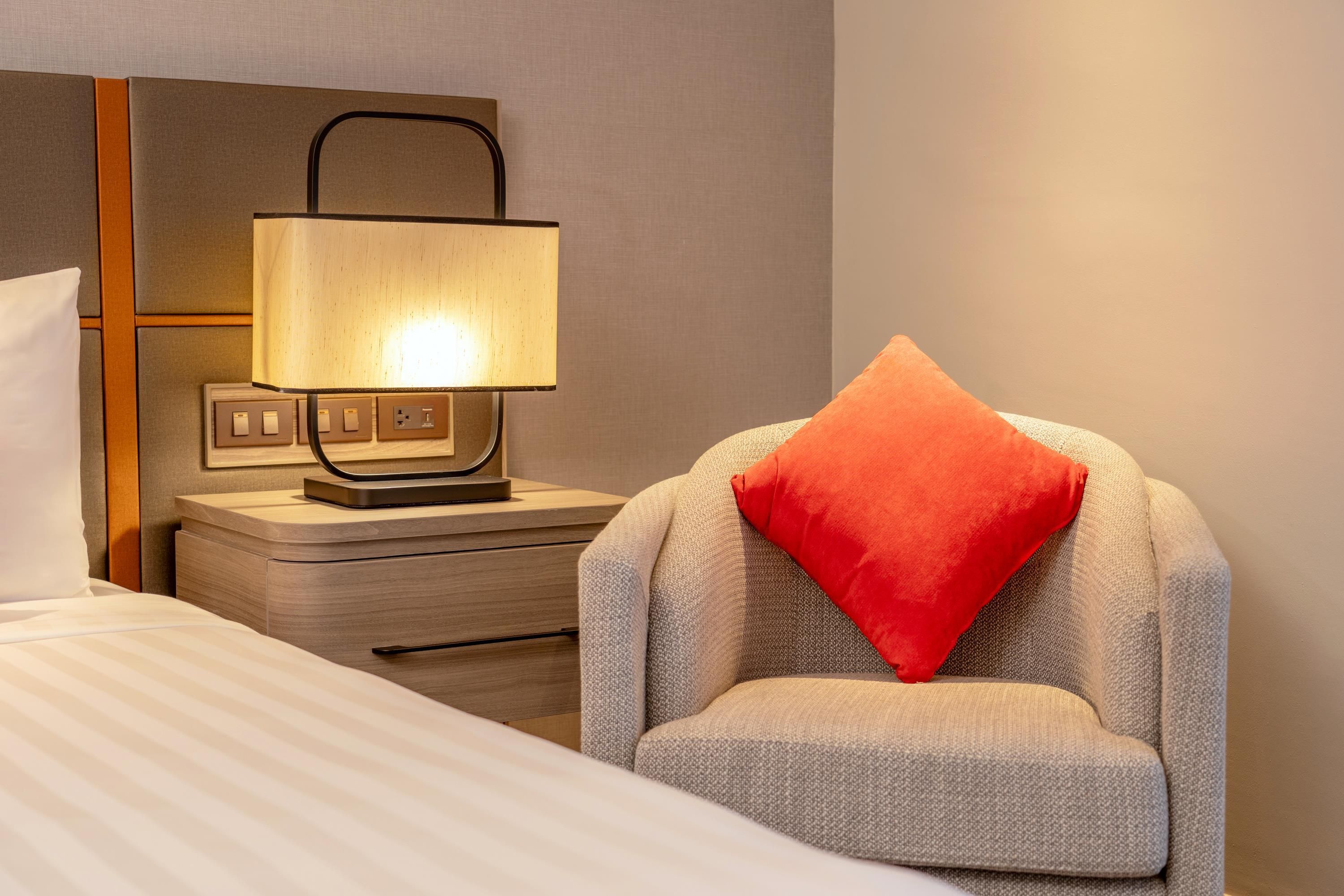 A hotel room with a bed, a nightstand with a lamp, and a chair with a pillow.