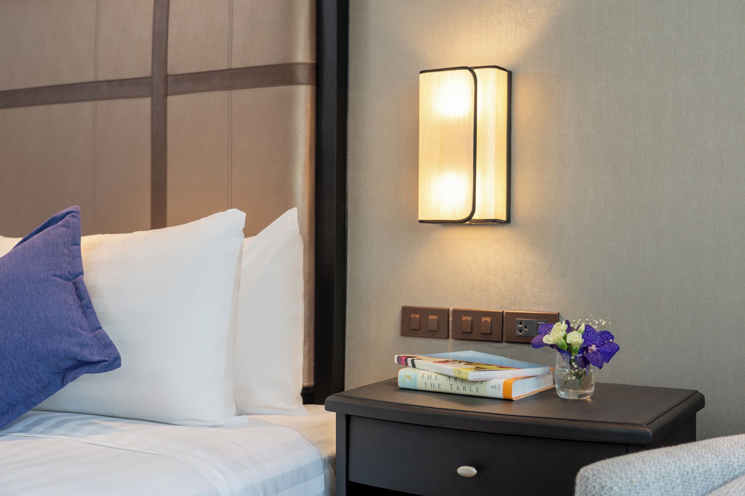 A neatly made bed with a blue pillow, a bedside table with books and a vase of flowers, and a wall-mounted light fixture.