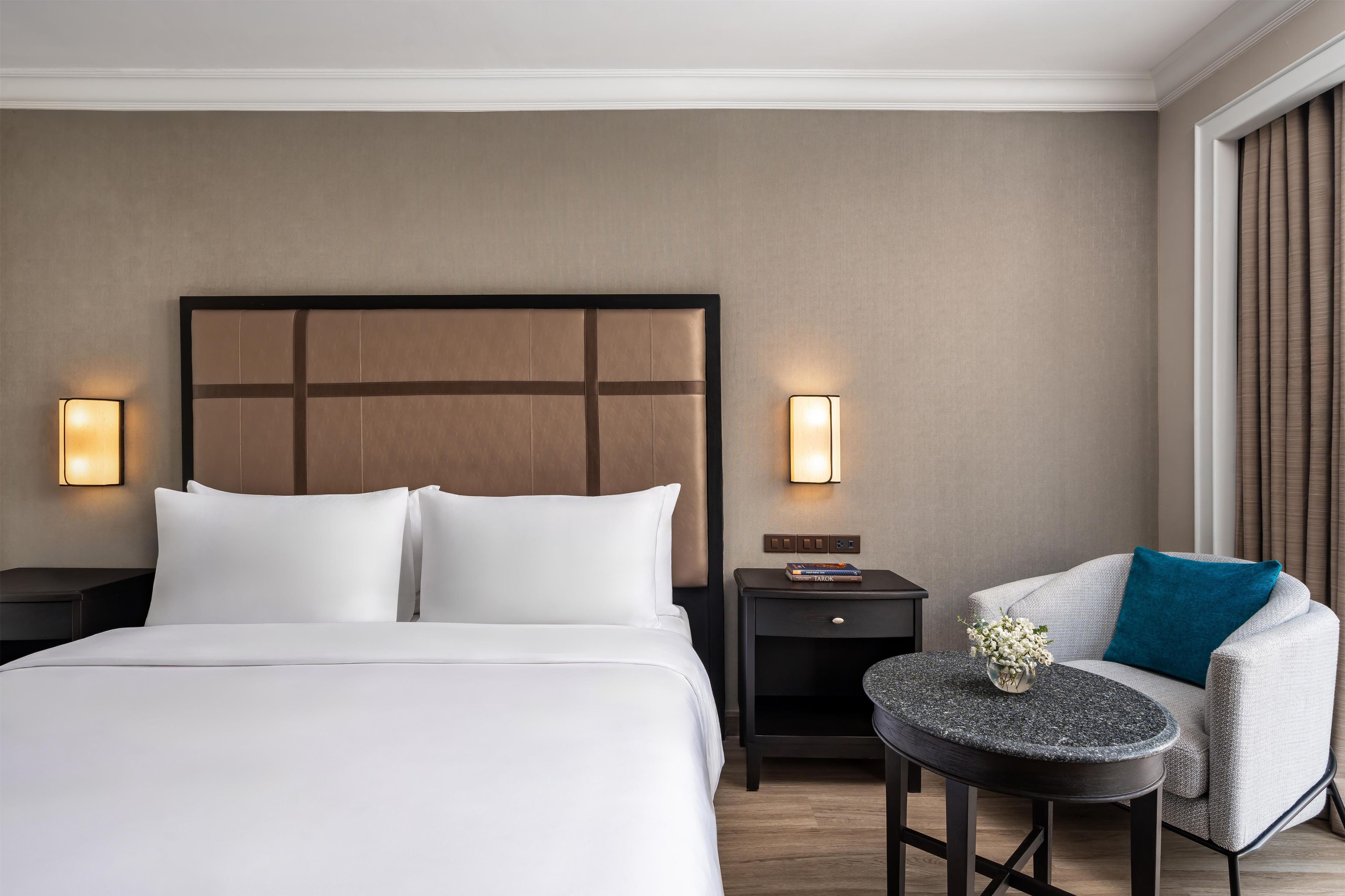 A hotel room with a large bed, two bedside tables with lamps, a small round table with a vase, and a chair with a cushion.