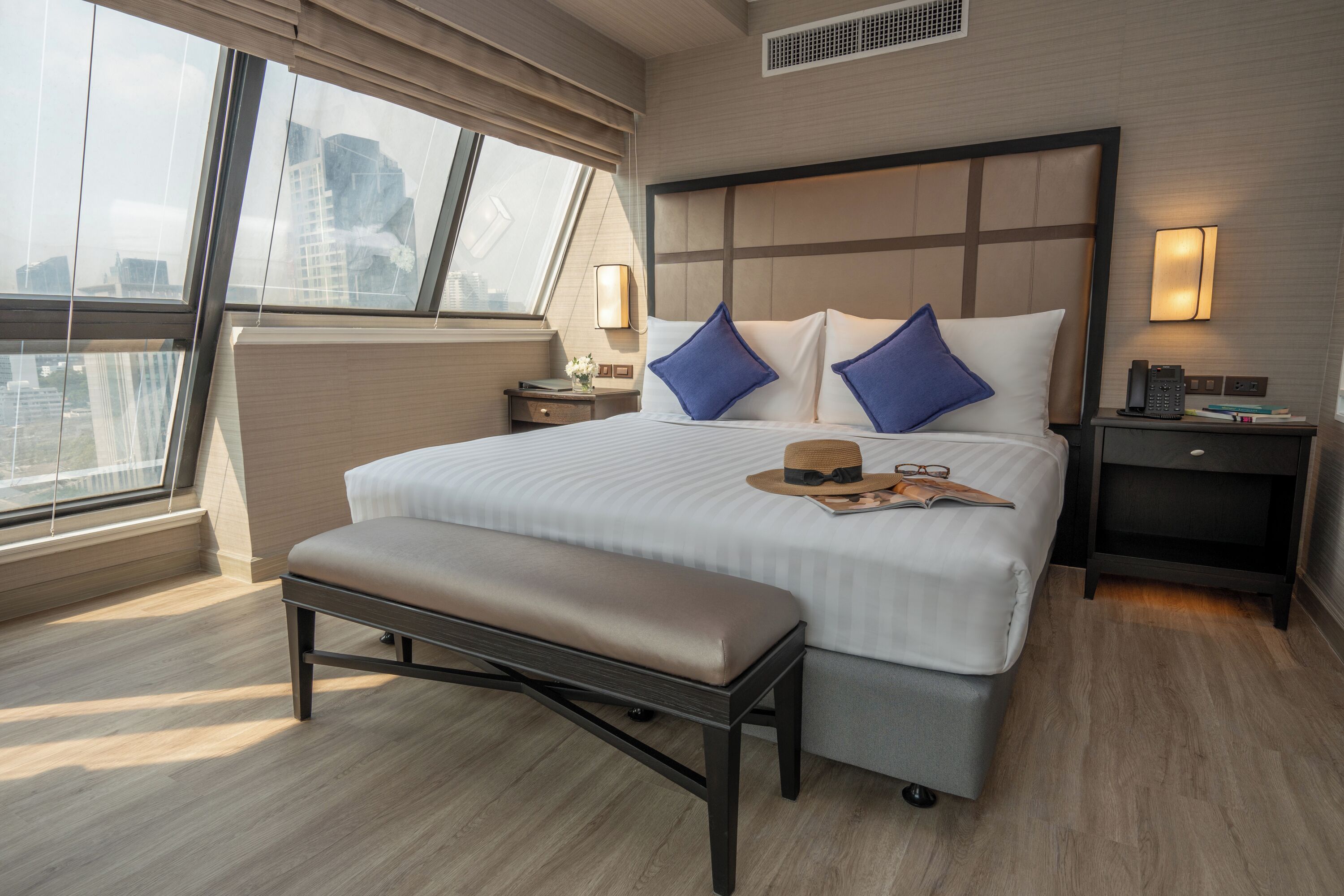 A modern hotel room with a large bed, a bench, a nightstand, and a window with a city view.