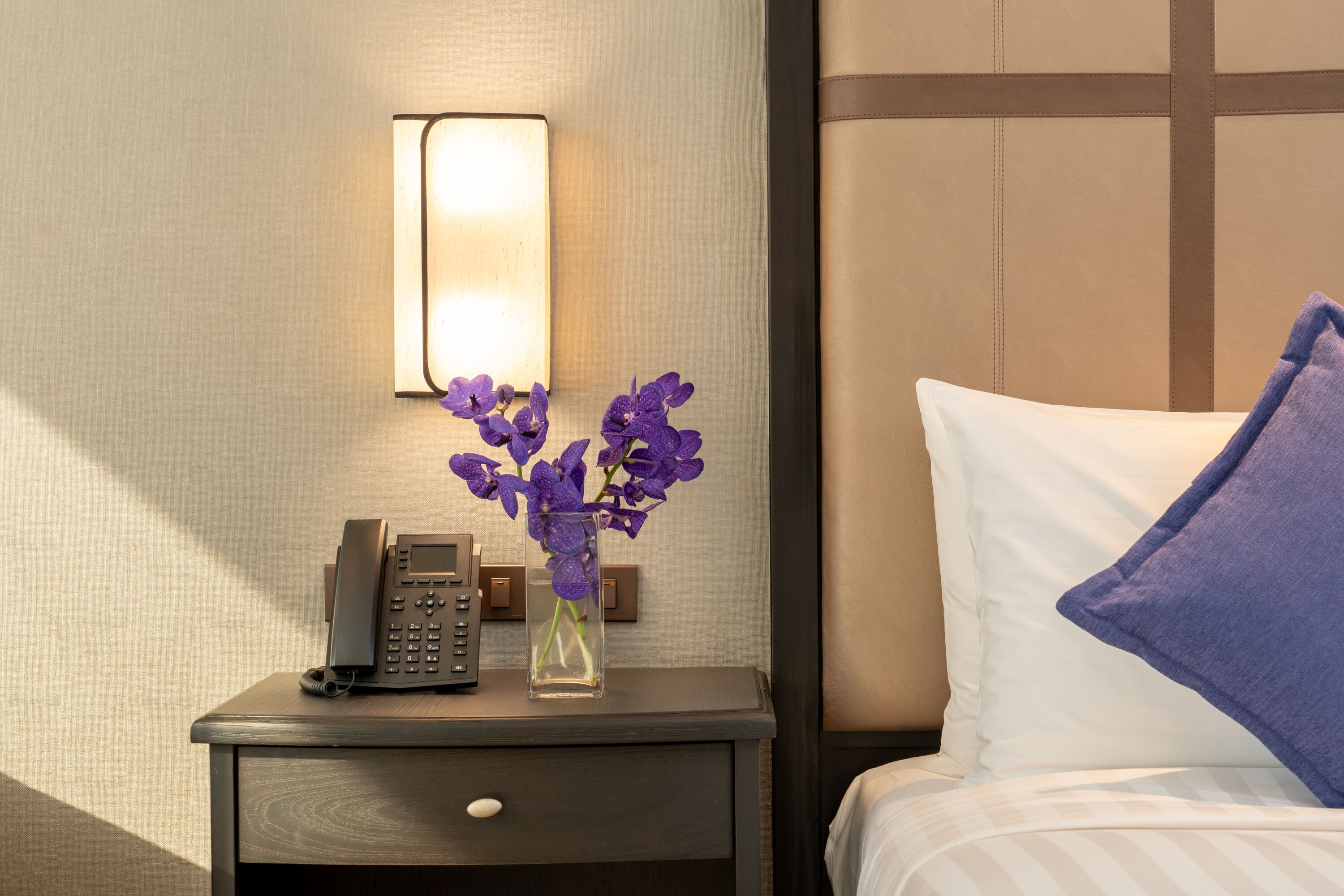 A neatly made hotel room with a bedside table, a phone, a vase with purple flowers, and a bed with a white pillow and a blue towel.