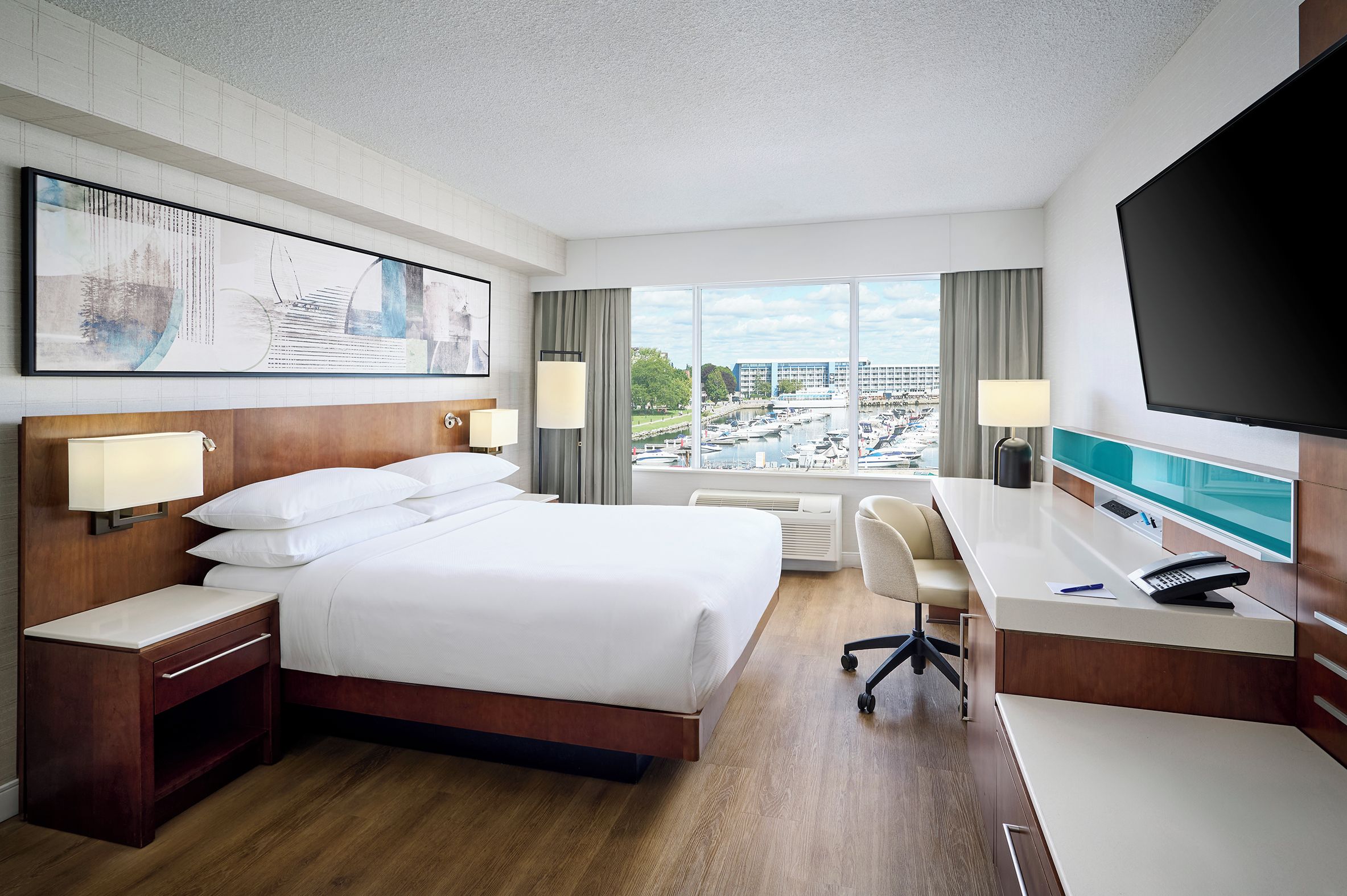 Een hotelkamer met een groot bed, een bureau, een stoel, een televisie en een raam met uitzicht op een jachthaven en gebouwen.