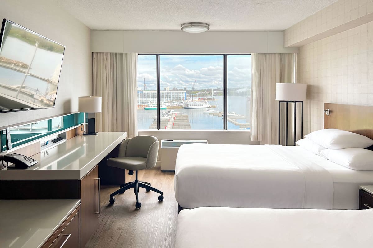 A hotel room with a bed, a desk with a chair, a TV, and a view of a harbor with boats.