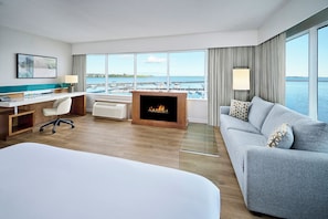 A hotel room with a bed, a desk, a fireplace, a sofa, and a large window with a view of the sea and boats. - Delta Hotels by Marriott Kingston Waterfront (Kingston)