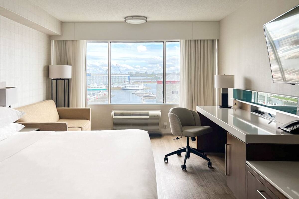 A hotel room with a bed, a desk, a chair, a TV, and a large window with a view of a marina and mountains.