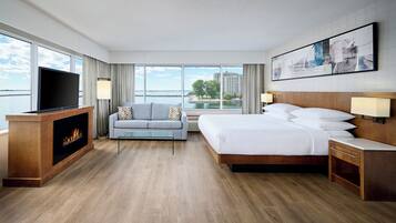A hotel room with a bed, a sofa, a TV, a fireplace, and large windows with a view.