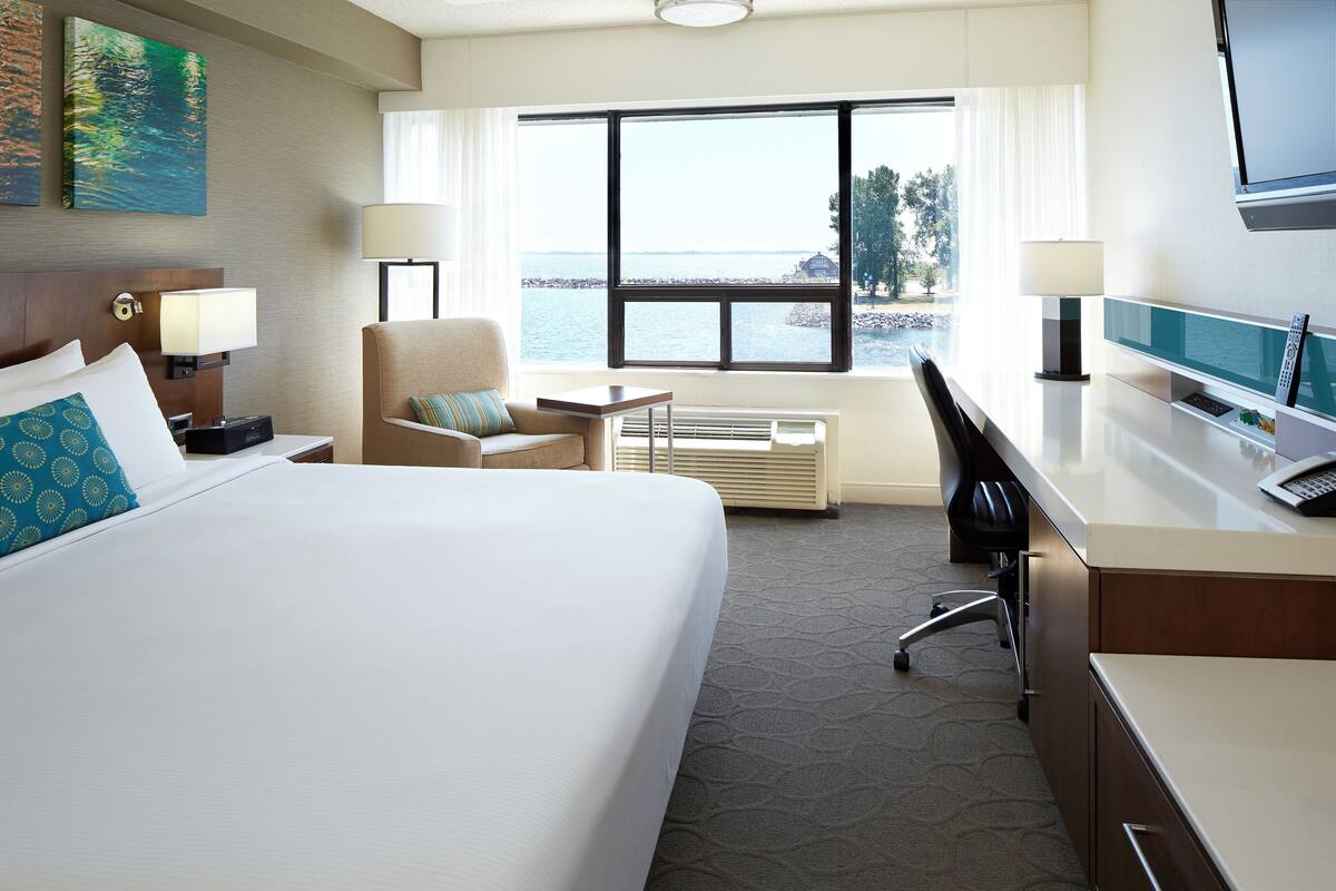 A hotel room with a large bed, a desk, a chair, a TV, and a window with a view of the water.