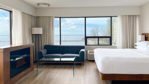 Habitación de hotel con una cama, un sofá, una mesa de centro de vidrio, un televisor y ventanas amplias con vistas al mar y edificios.