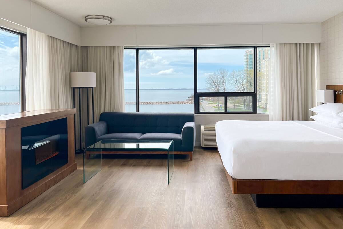 A hotel room with a bed, a sofa, a glass coffee table, a TV, and large windows with a view of the sea and buildings.