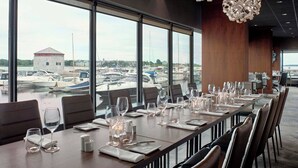 Un comedor con vista al puerto deportivo, que cuenta con una mesa larga dispuesta con vasos y platos, y una lámpara de araña moderna.