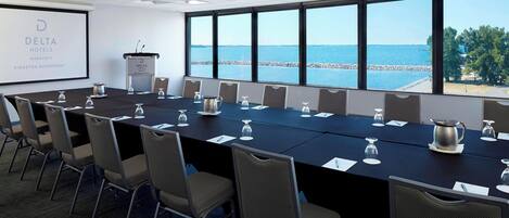 A conference room with a long table, chairs, a projector screen, and a view of the sea.