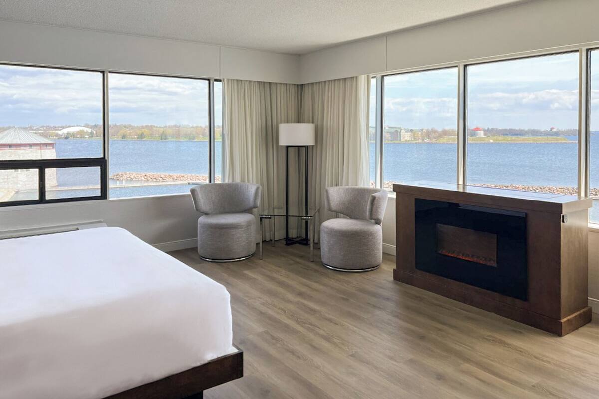 A hotel room with a bed, two armchairs, a fireplace, and large windows overlooking a body of water.