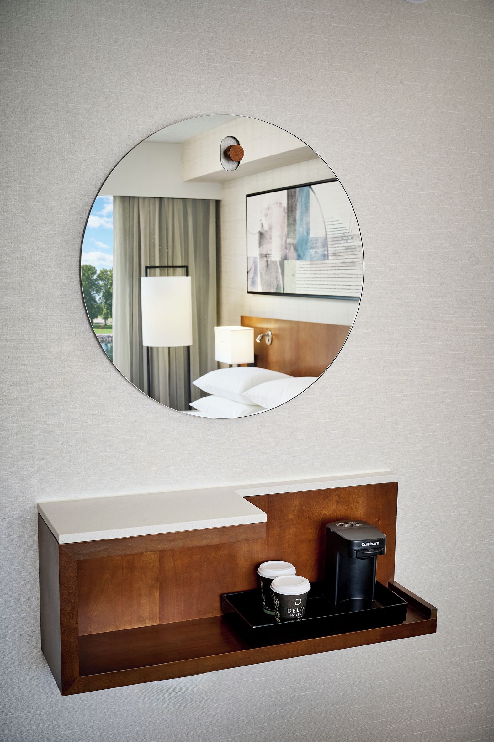 Une salle de bain d’hôtel avec un miroir rond, un meuble-lavabo en bois, une machine à café et un plateau avec deux tasses.