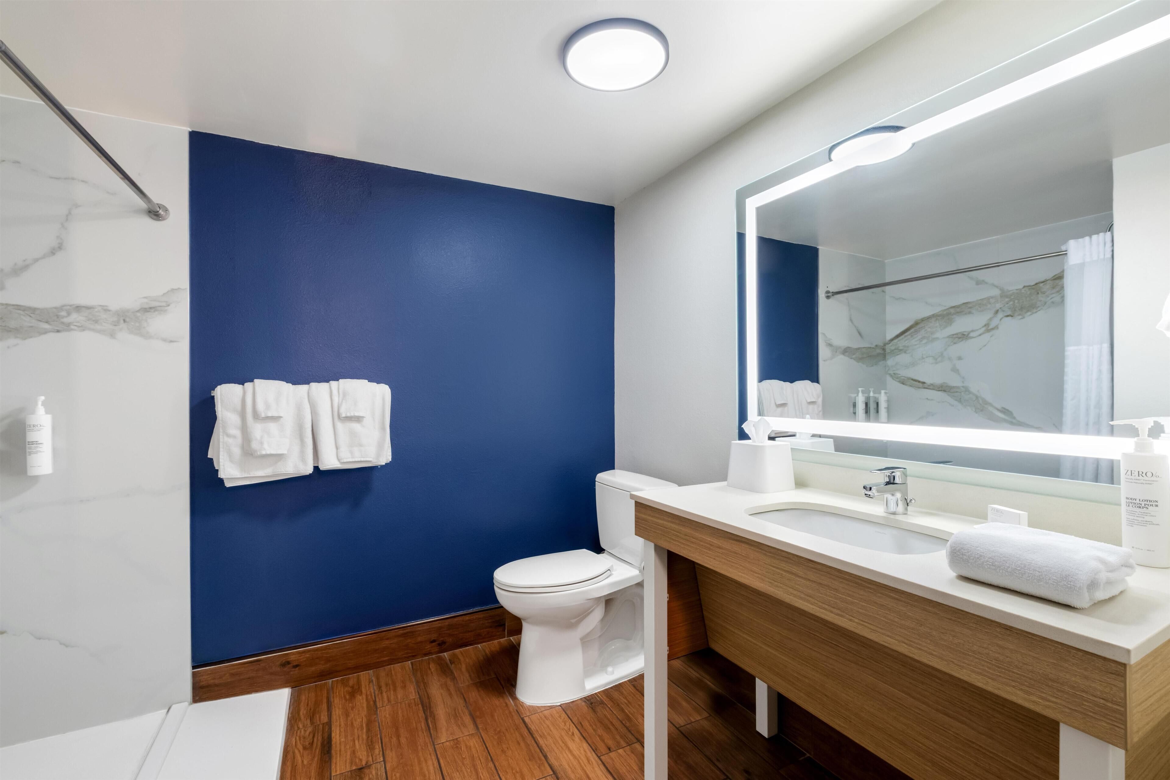 A modern bathroom with a blue accent wall, a white toilet, a wooden vanity, and a large mirror.