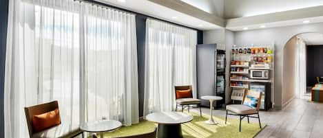 Un hall d’hôtel moderne doté de grandes fenêtres, d’espaces aménagés pour s’asseoir, d’un réfrigérateur et d’un kiosque en libre-service.