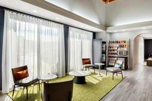 A modern hotel lobby with large windows, seating areas, a refrigerator, and a self-service kiosk.
