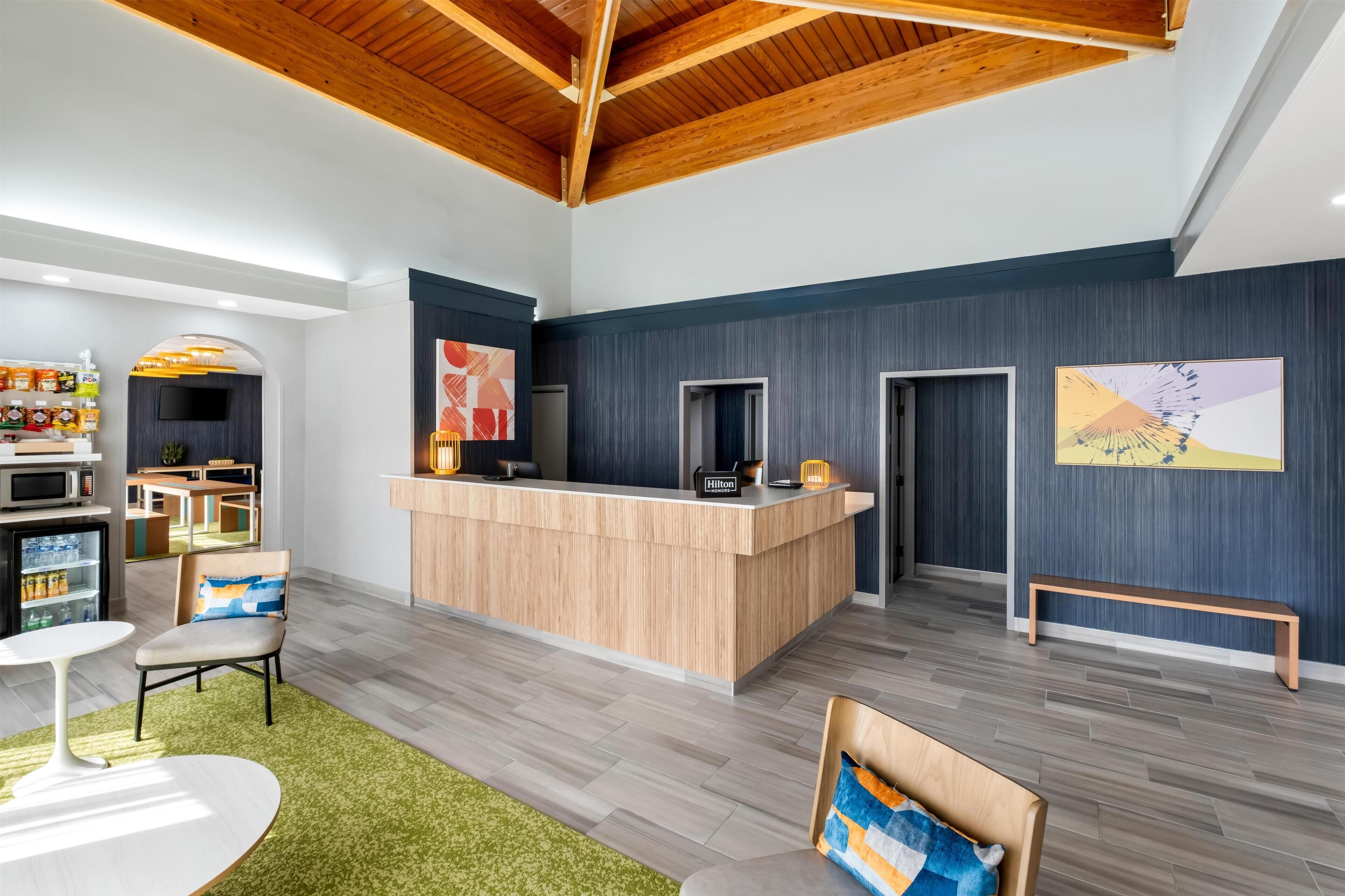 A modern hotel lobby with a wooden reception desk, seating area, and a kitchenette.