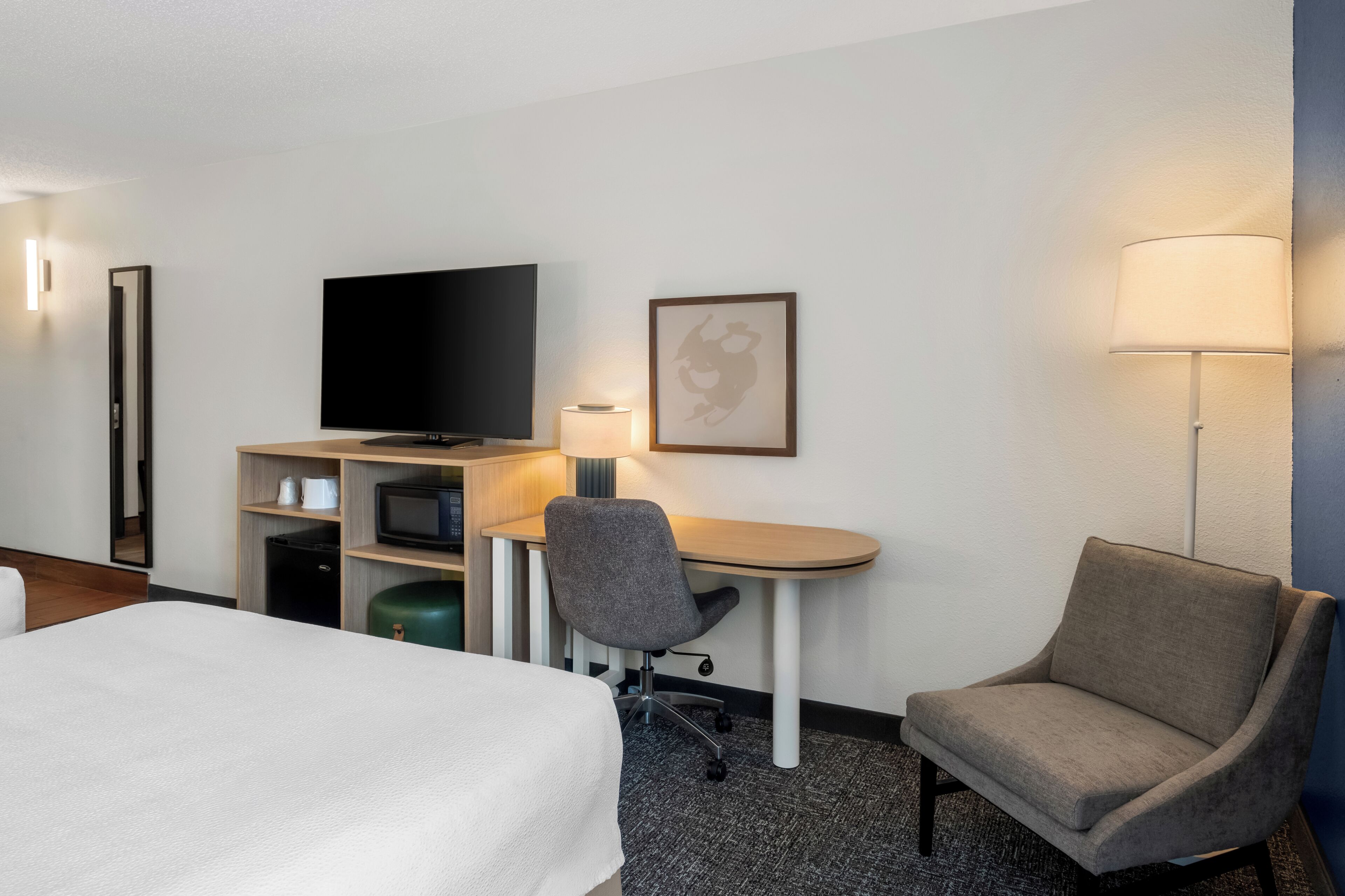 A hotel room with a bed, a desk with a chair, a television, a lamp, and a framed picture on the wall.
