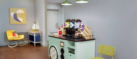 A room with a teal-colored kitchenette, two yellow chairs, a small table with a lamp, and a wall-mounted abstract art piece.