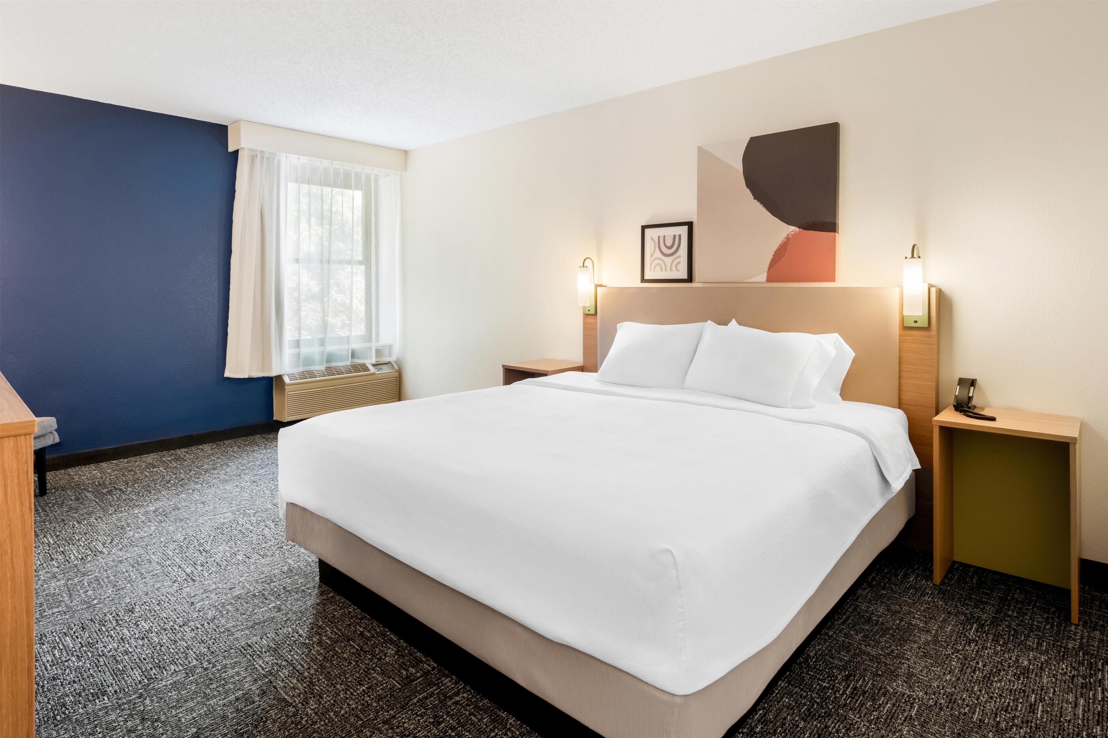 A hotel room with a large bed, bedside tables, a window with curtains, a wall-mounted artwork, and a carpeted floor.