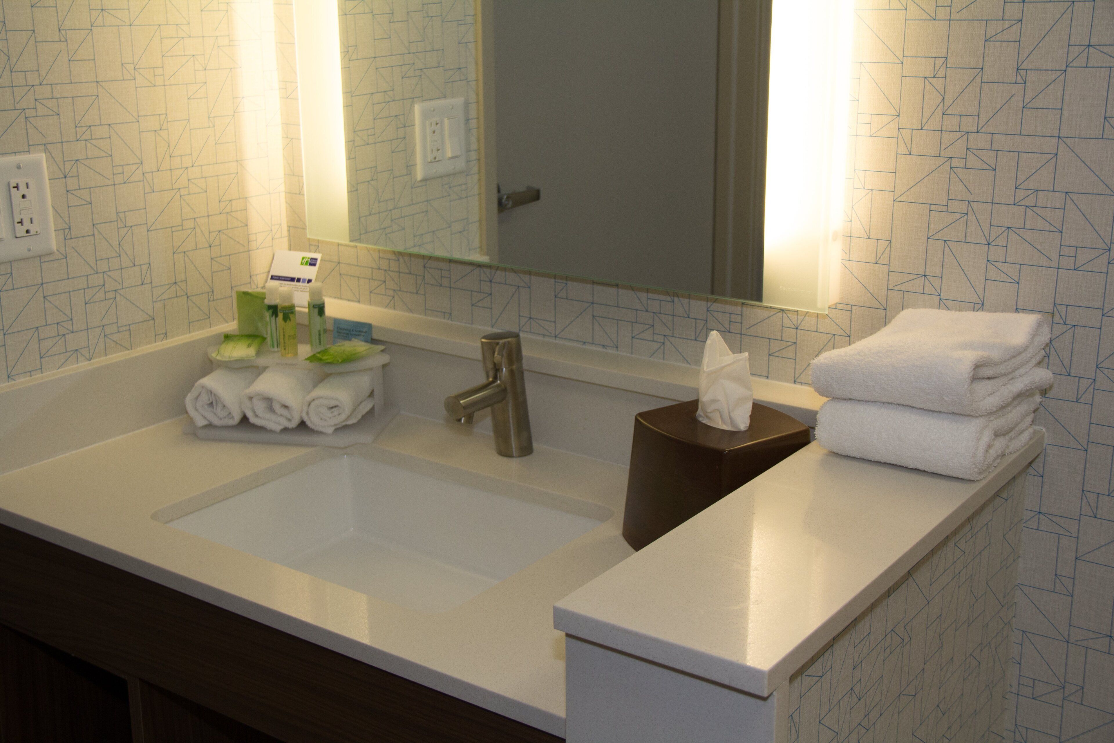 A hotel bathroom with a sink, mirror, towel dispenser, and toiletries.