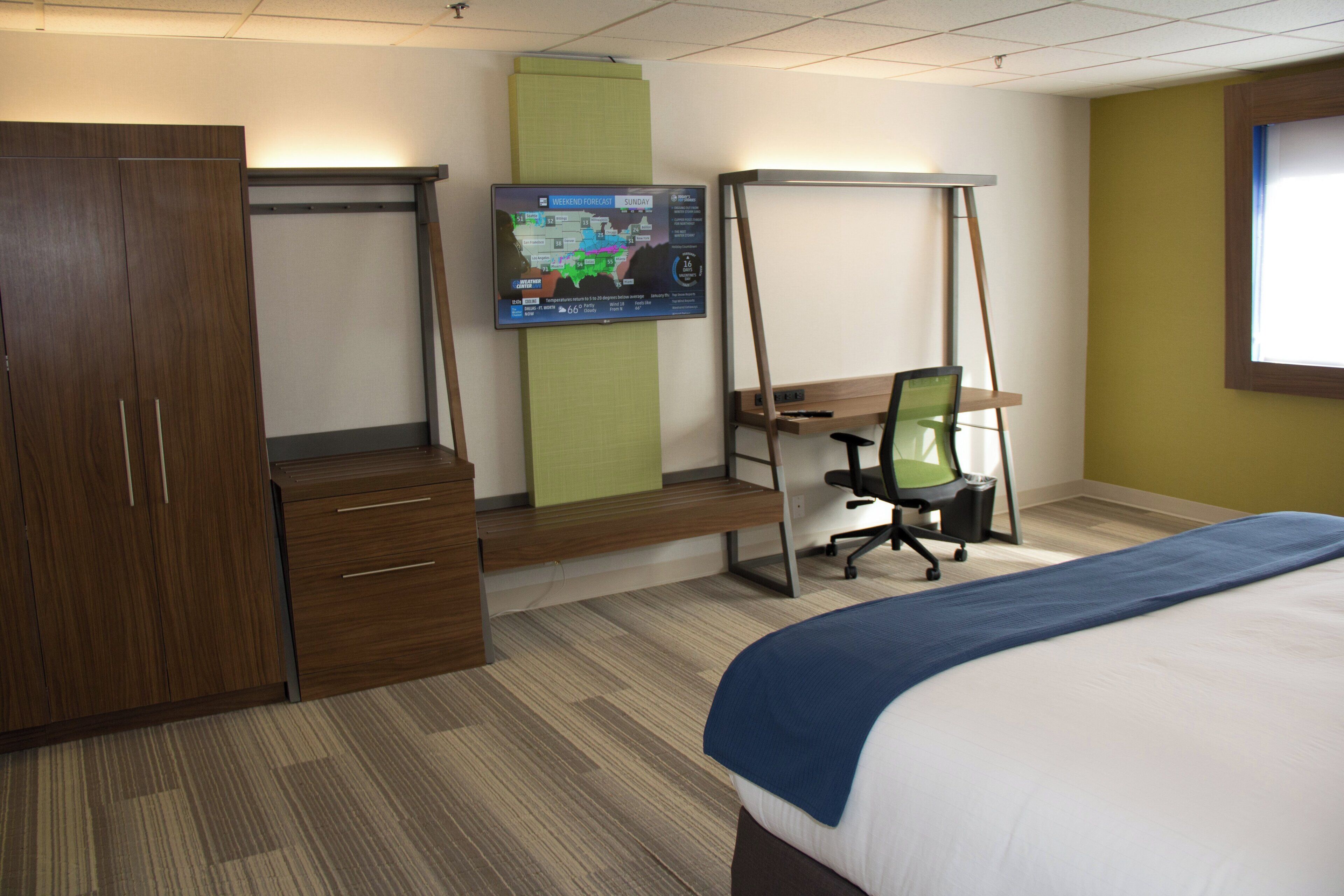 Ein Hotelzimmer mit Bett, Schreibtisch, Stuhl, Kleiderschrank und einem an der Wand befestigten Fernseher, der eine Wettervorhersage zeigt.