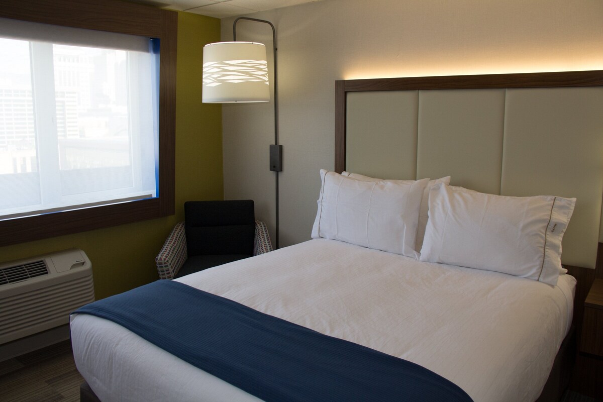 Une chambre d’hôtel avec un grand lit, une table de chevet, une lampe et une fenêtre avec des stores.