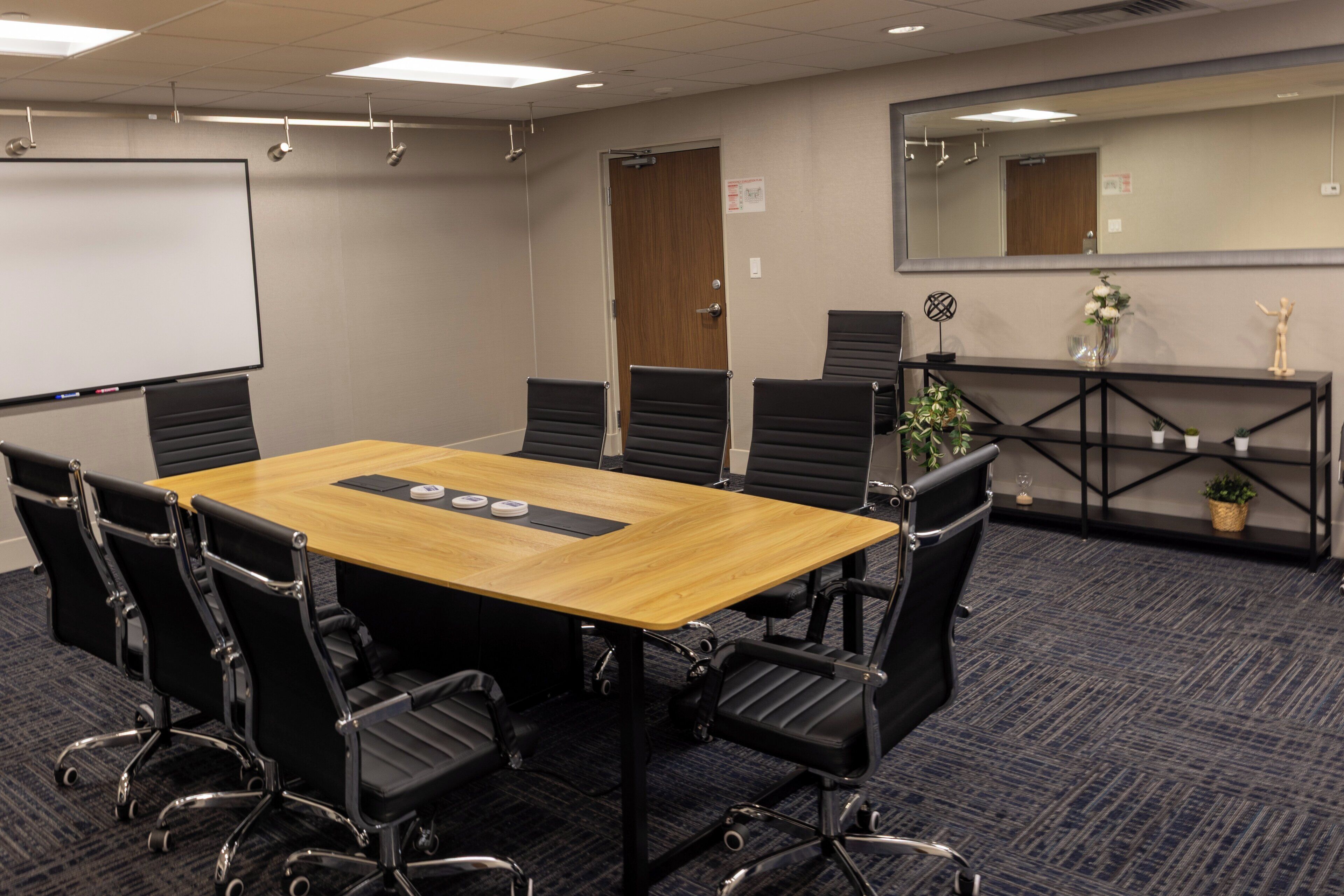 A conference room with a wooden table, black chairs, a whiteboard, and a mirror on the wall.