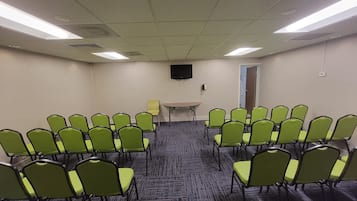 Un salón de conferencias con filas de sillas verdes, una mesa pequeña, un televisor de pantalla plana y una puerta.