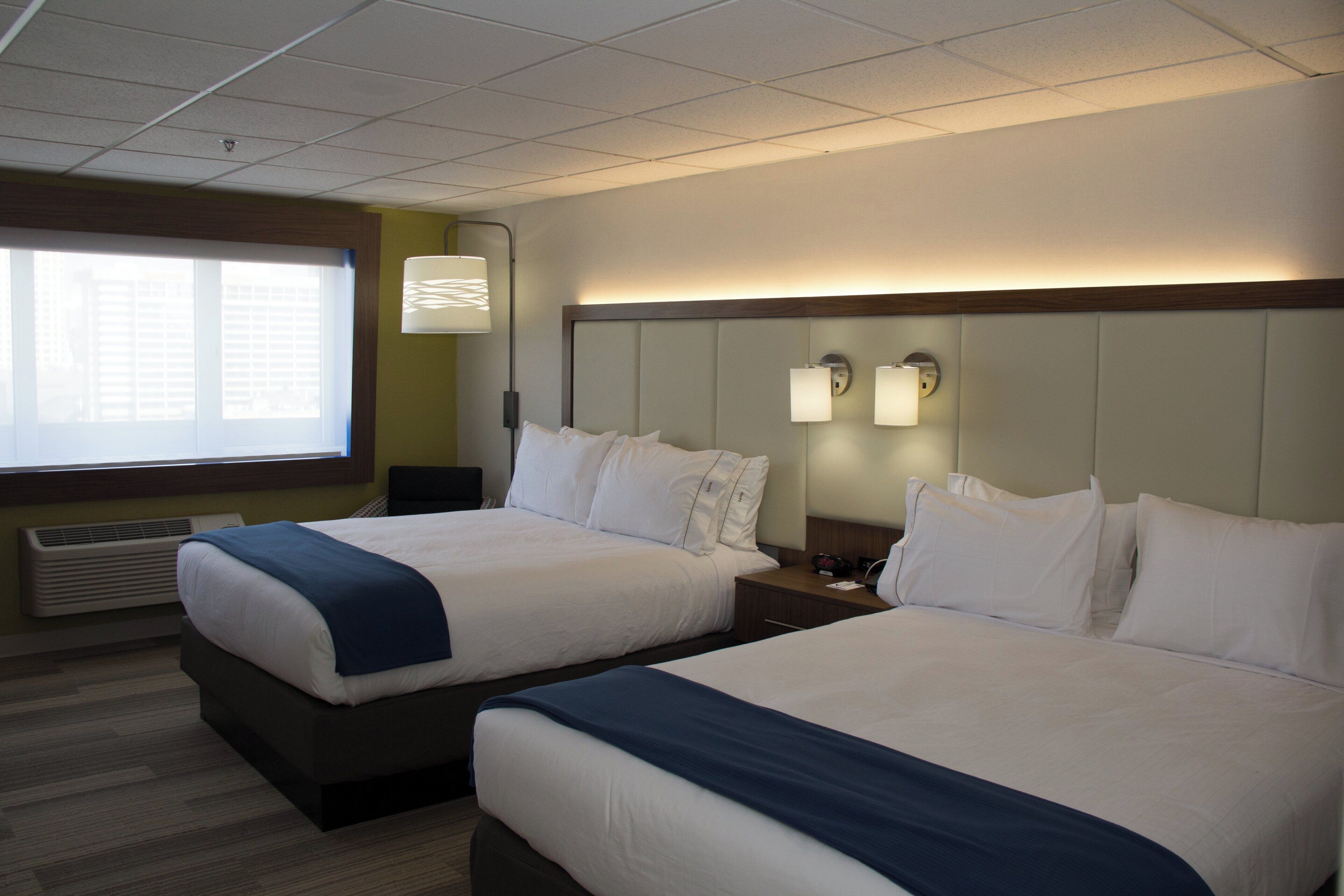A hotel room with two beds, a large window with blinds, a wall-mounted lamp, and a clock.