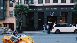A Tilden Hotel with a sign for Tails & Kitchen, a white car parked in front, and a yellow taxi with passengers. - Tilden Hotel (San Francisco)