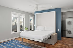 Un dormitorio moderno con cama, ventilador de techo, ventana con vista a la ciudad y lavadora en una esquina.