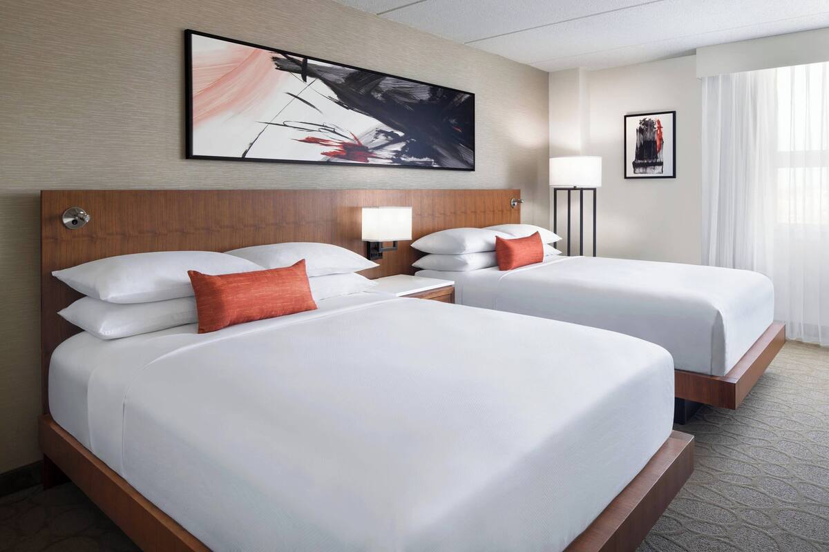A hotel room with two beds, a wooden headboard, a large abstract painting, a floor lamp, and a window with curtains.