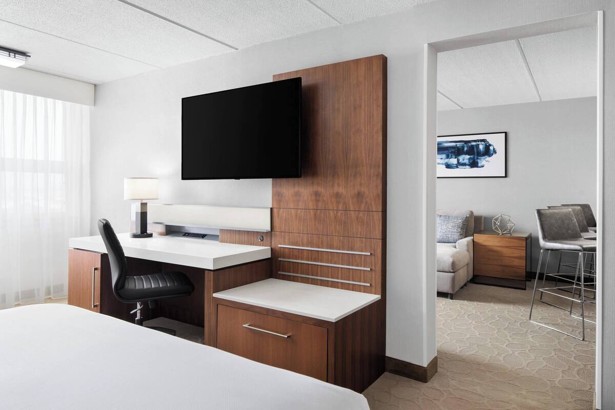 A hotel room with a bed, a desk with a chair, a television mounted on a wooden panel, a small table with a chair, and a framed picture on the wall.