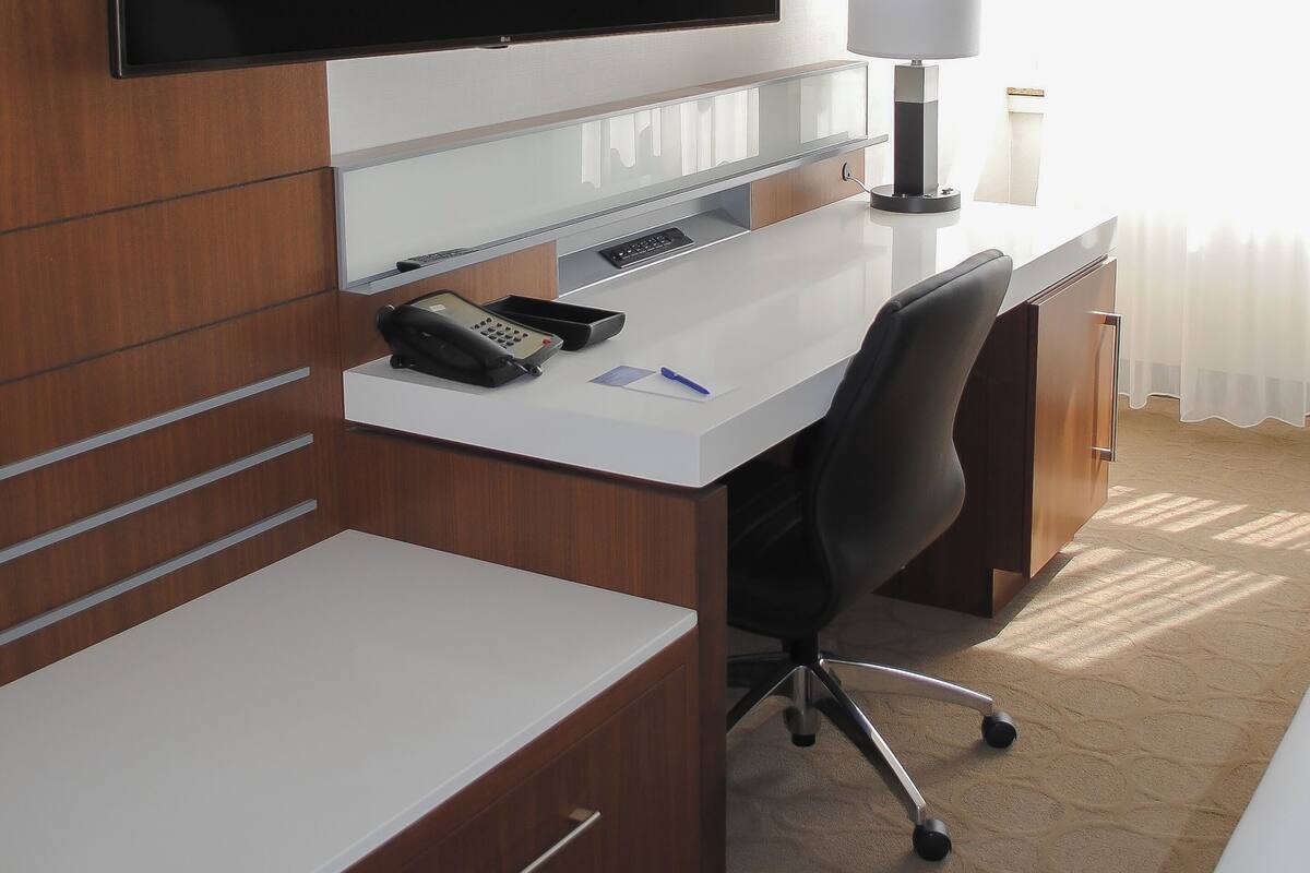 A hotel room with a large flat-screen TV mounted on the wall, a desk with a telephone, a lamp, and a chair.