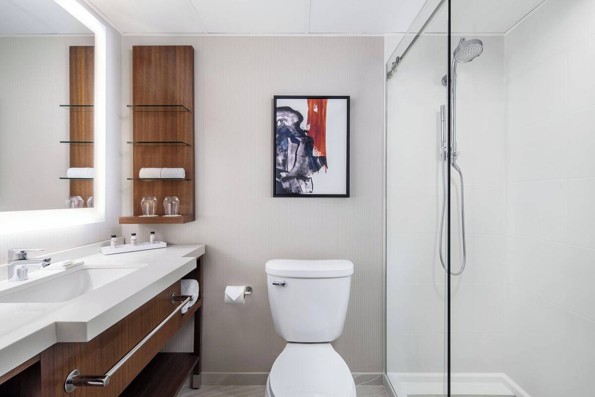 A modern bathroom with a white sink, wooden vanity, a framed abstract painting, a toilet, and a glass-enclosed shower.