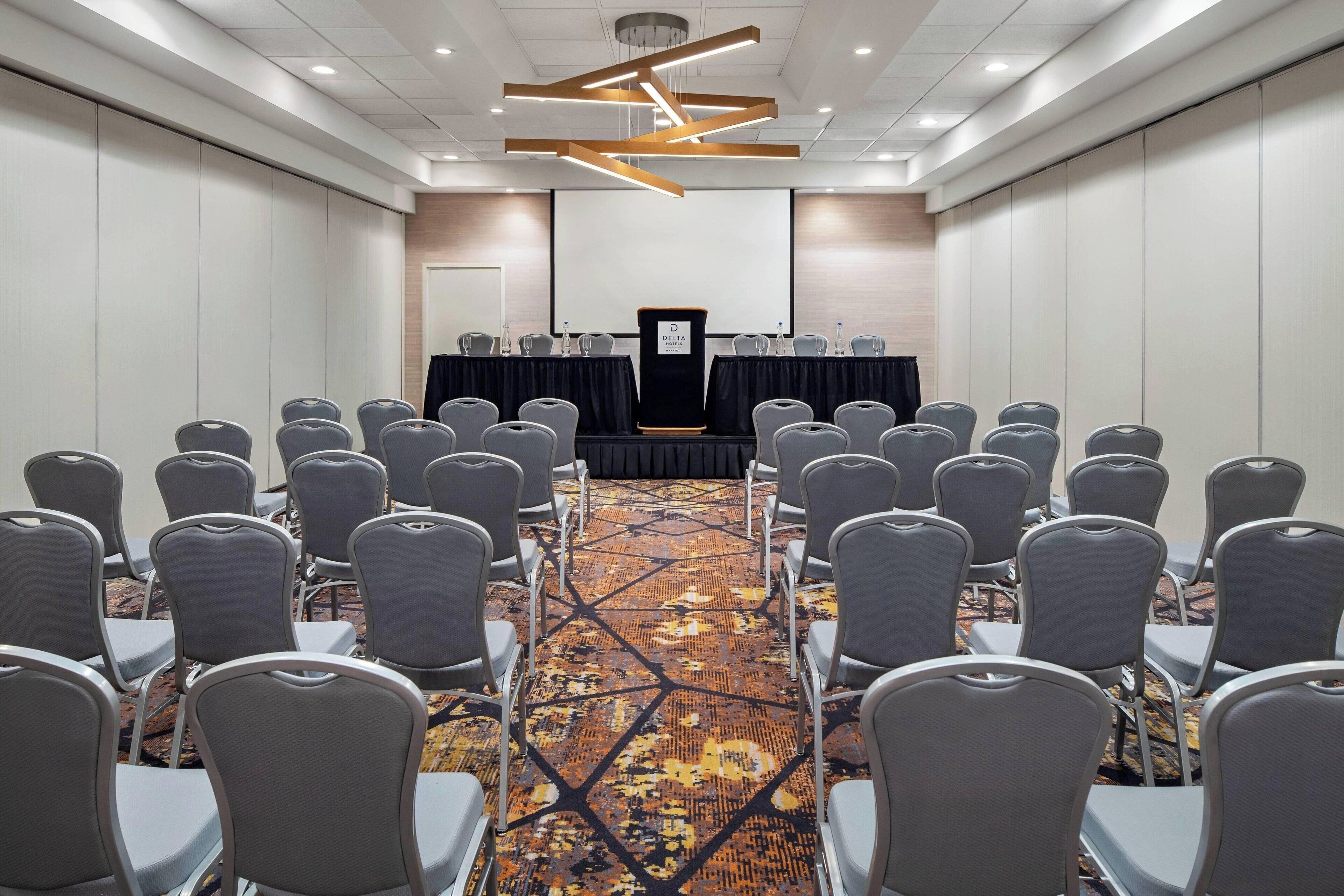 A conference room with rows of chairs facing a podium, a carpeted floor, and a modern ceiling design.